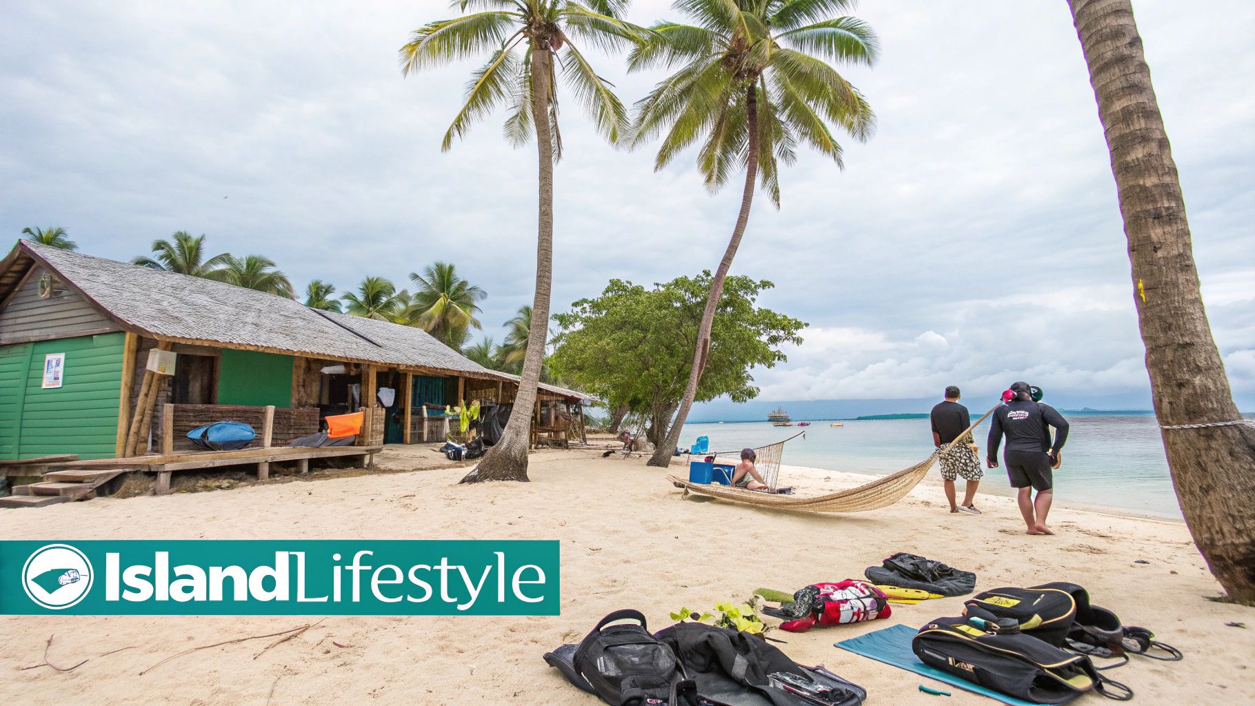 A tropical island beach with wooden bungalows, palm trees, people, and gear by the ocean.